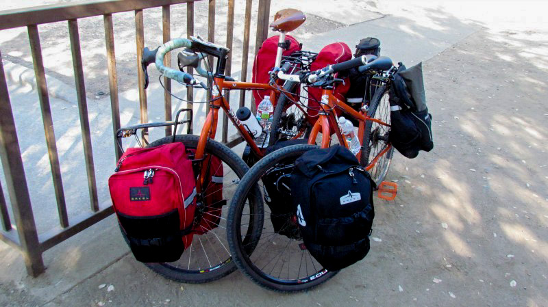 Front view of 2, orange Surly bikes loaded with gear, parked side by side, next to a handrail with snow behind it
