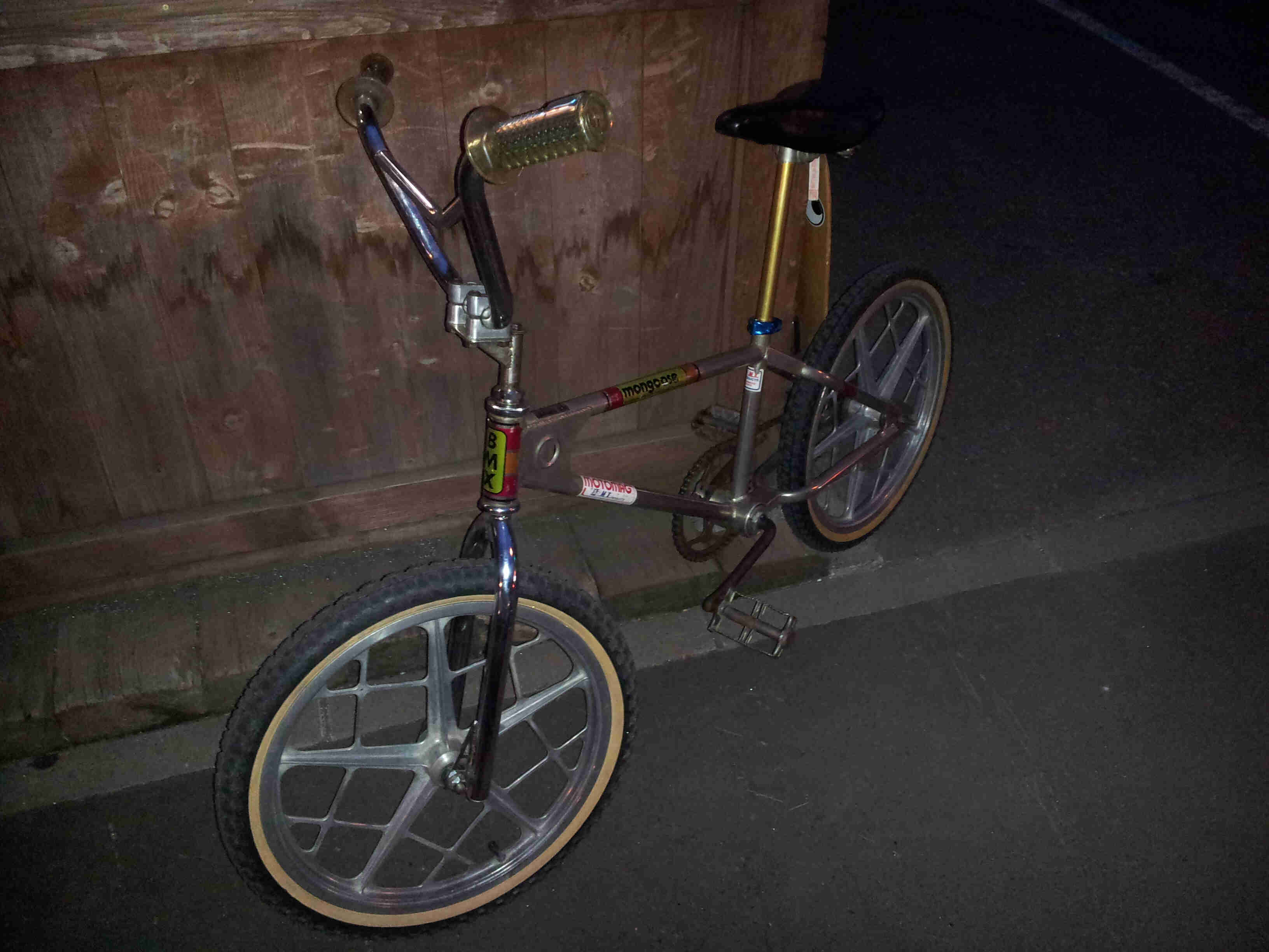 Left side, dimly lit view of a chrome BMX bike, leaning against a wood wall