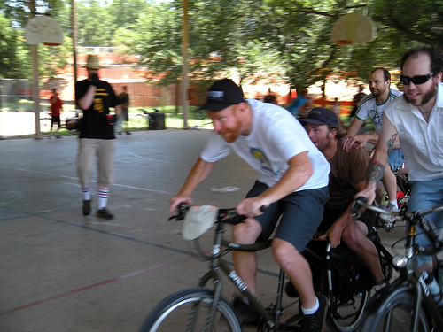 Front, left side view of cyclists, on Surly Big Dummy bikes with people on back, racing on an outdoor basketball court