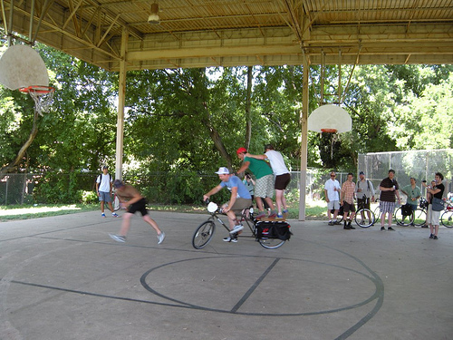 Left side view of a cyclist, riding a Surly Big Dummy bike with 2 people standing on back, on a outdoor basketball court