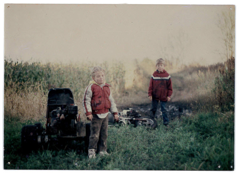 Front view of 2 children, standing between 2 cornfields, next to a go-cart