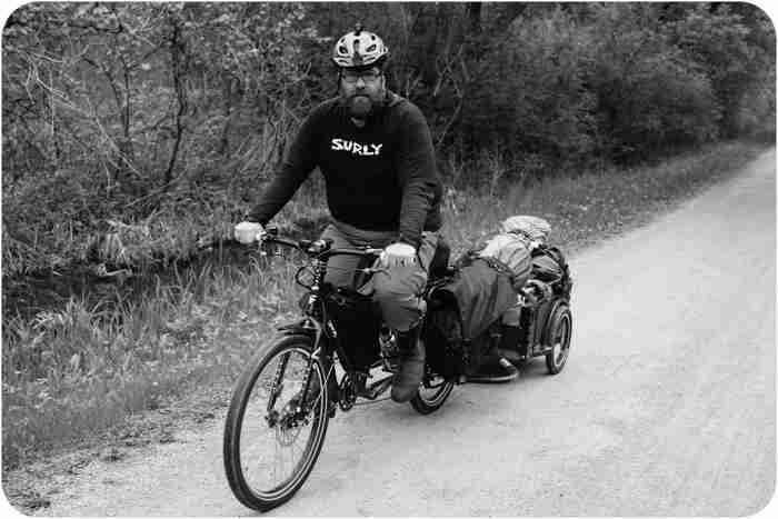 Front, left side view of a cyclist, riding a Surly bike with gear and trailer, on a gravel road - black & white image
