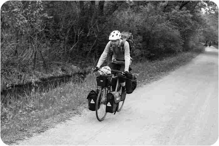 Front, left side view of a cyclist, riding a Surly bike with gear, on a gravel road, along a stream - black & white