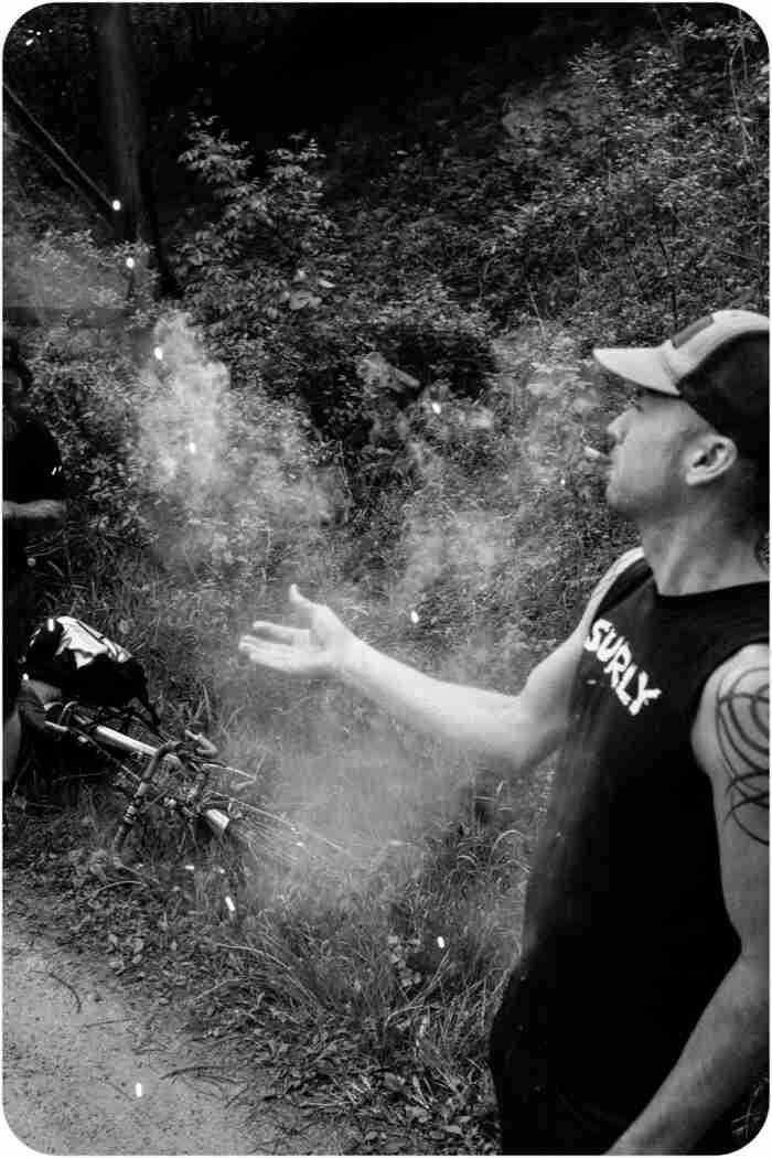 Left side view of person wearing a Surly shirt, with smoke around their right hand, in the woods - black & white image