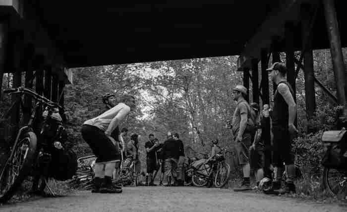 Side view of cyclists, standing with their bikes, on a gravel road under bridge in the woods - black & white image