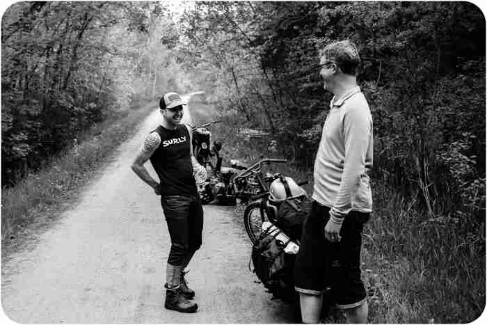 Front view of a person wearing a Surly shirt, standing on a gravel road next to another person and bikes - black & white