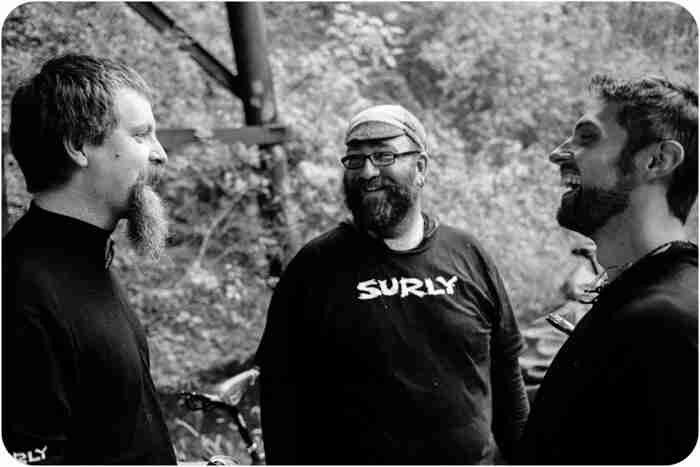 Front view of a person wearing a Surly shirt, standing between 2 other people - black & white image