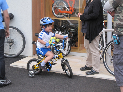 Right side view of a small child, riding a BMX bike with training wheels, inside of a bike shop with people around