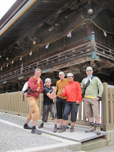 Front view of 5 people posing for a picture, standing on a walkway in front of a Japanese building