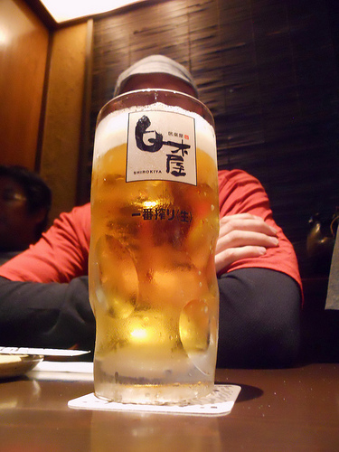 Front view of a mug of beer, sitting on a table, covering the face of a person sitting behind it, inside of a restaurant