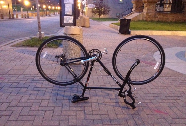 Right side view of a black Surly bike, flipped upside down and standing on the seat and handlebar, on a brick sidewalk
