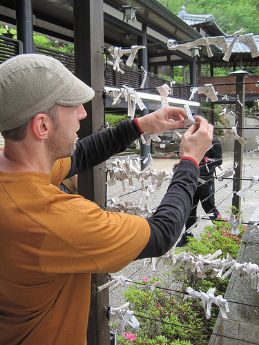 Rear, right side view of a person, wrapping a small piece of paper around a fence wire, in a Japanese garden
