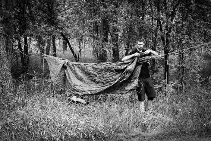 Front view of a person hanging a tarp over a rope tied between 2 trees, in the woods - black & white image