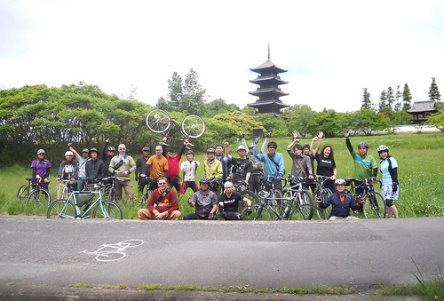 Front view of a group of cyclists and bikes, lined up side by side in front of a road, with a field and pagoda behind