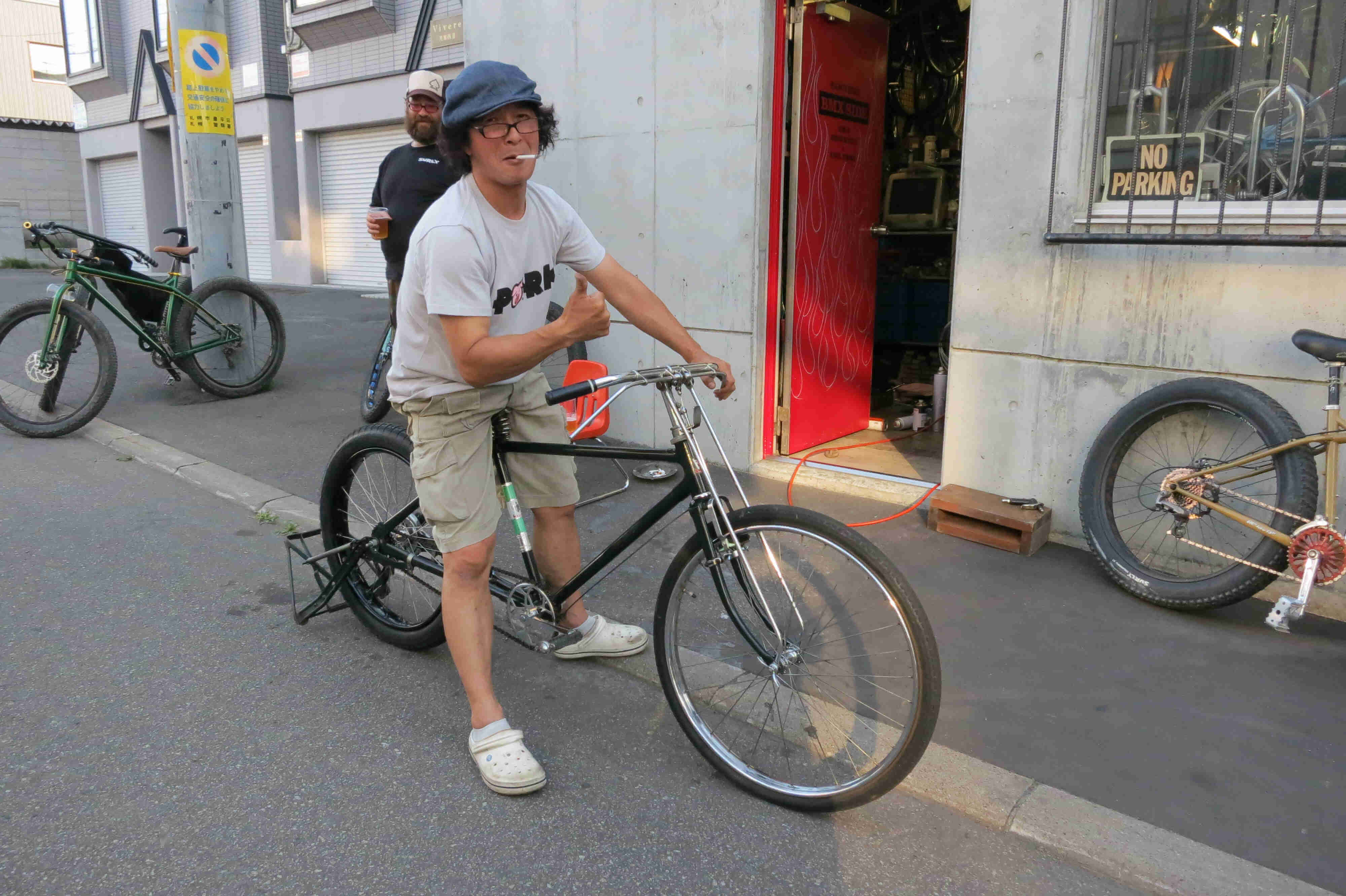 Right side view of a cyclist, sitting on a bike with a cigarette in their mouth, on a sidewalk in front of a bike shop