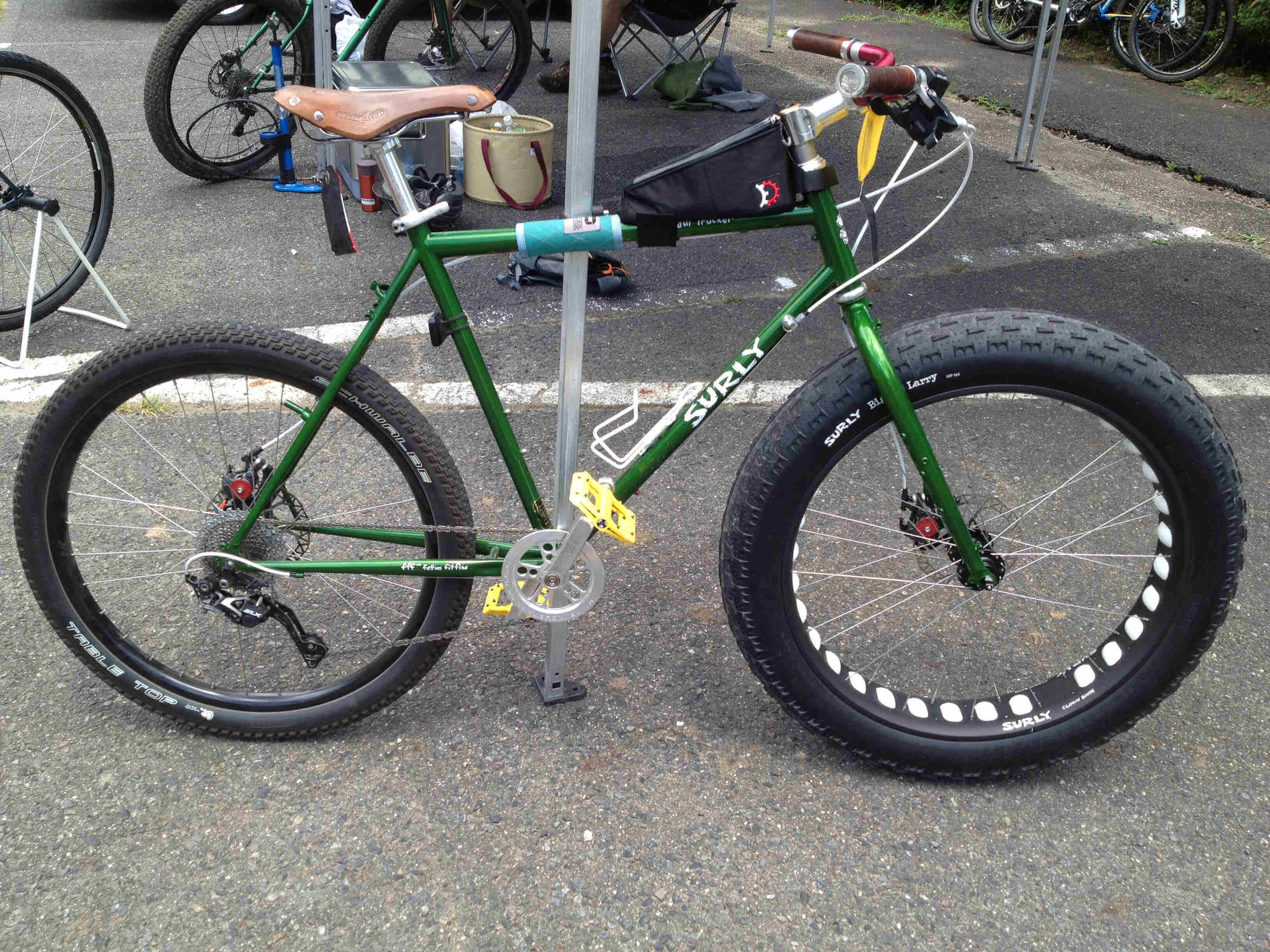 Right side view of a green Surly Long Haul Trucker bike, with a fat wheel on the front, leaning on a canopy pole