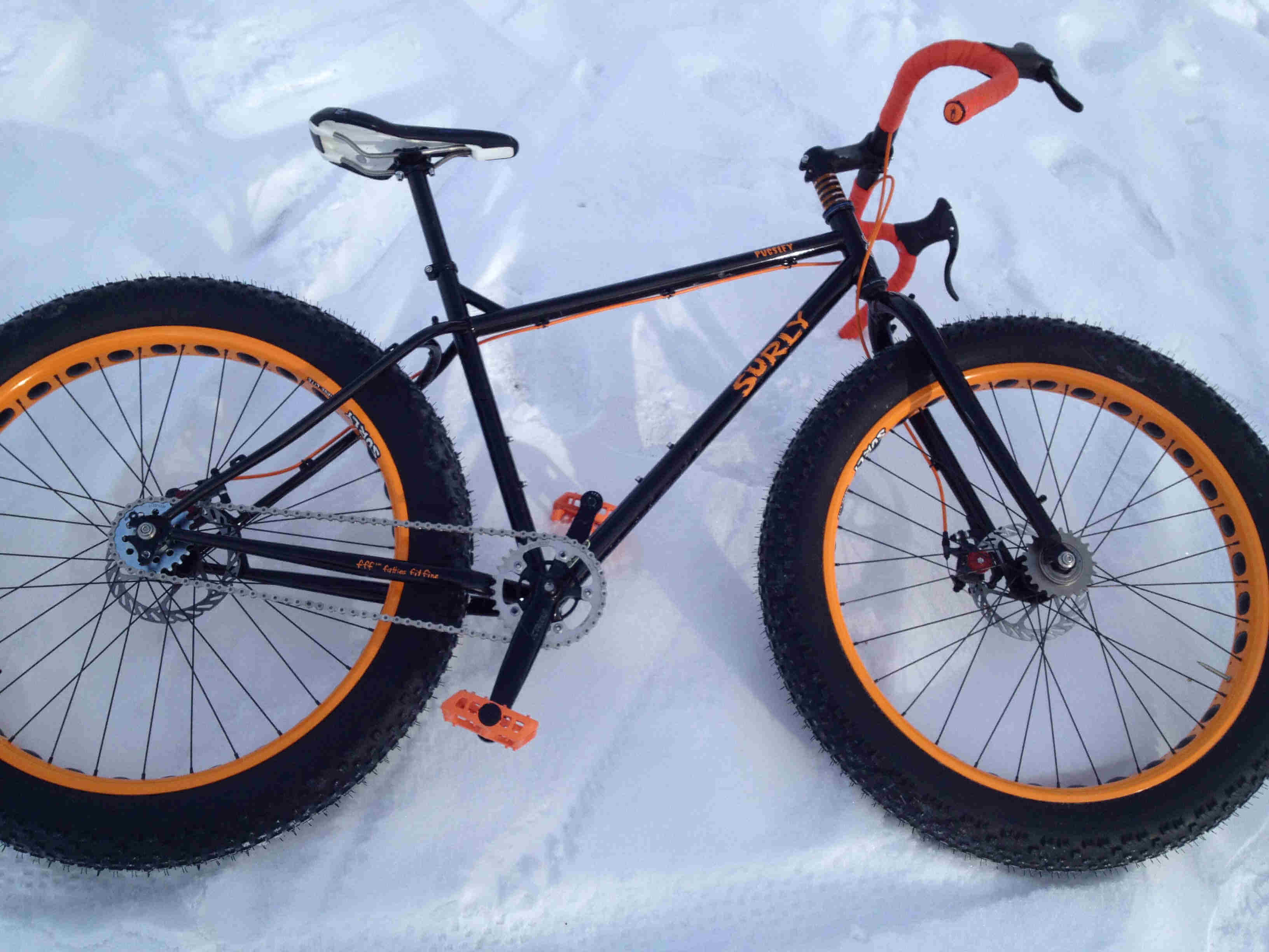 Downward, right side view of a black Surly Pugsley bike with orange rims, laying on it's left side in snow