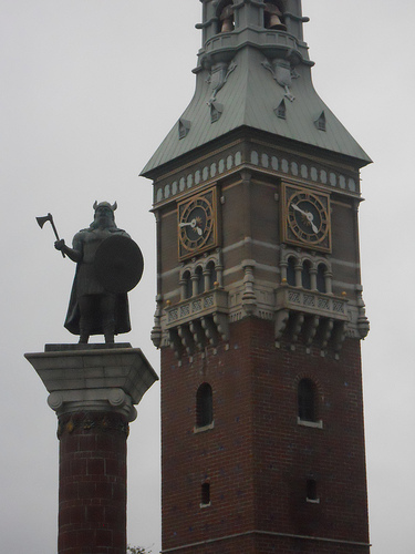 A clock tower next to a monument with the statue of a warrior on top