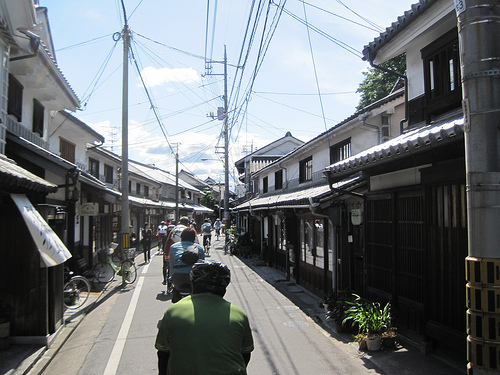 A rear view of cyclists, riding their Surly bikes straight away on a narrow paved street, with buildings on both sides