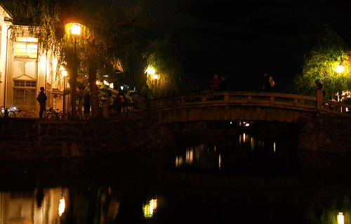 Side view of a small bridge with people walking, spanning over a river, at night with street light on the sides