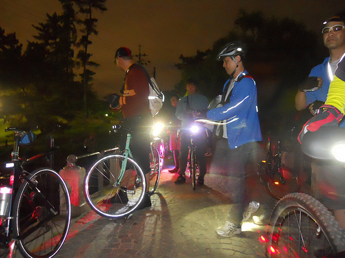 A group of cyclists and their bikes with headlights on, standing on a dirt road at night, with trees on the sides