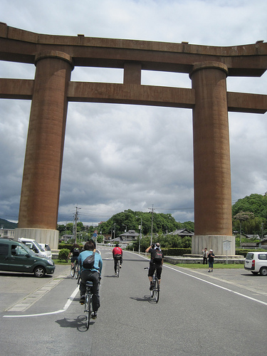 Rear view of a group of cyclists, riding their bikes toward a large Japanese torii that is arching over the street