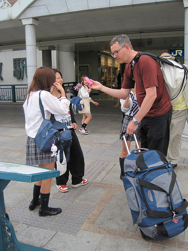 Right side view of a person showing their pink cell phone to 2 school children, on a sidewalk in front of a building