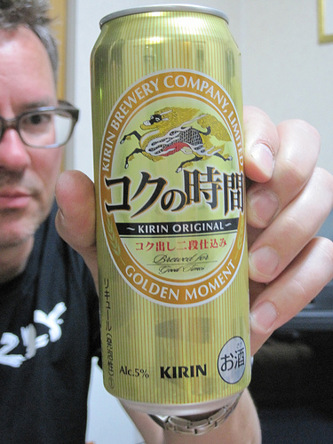 Front view of a Japanese beer can being held up by a person behind, inside of a room with white walls