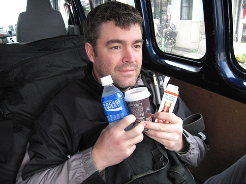 Front, chest up view of a person, sitting inside of a van, holding drinks up underneath their chin
