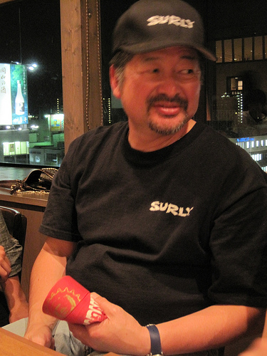Front view of a person sitting in a chair, wearing a black Surly logo hat & t-shirt, at night with building behind them