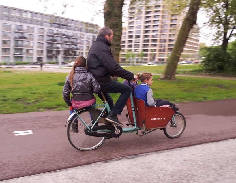 Right side view of a cyclist, riding a bike with a child sitting on the back rack, and another in a front bin