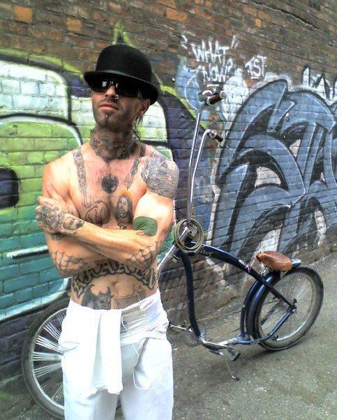 Front view of a person wearing a derby, in front of a chopper bicycle with a graffitied wall behind it