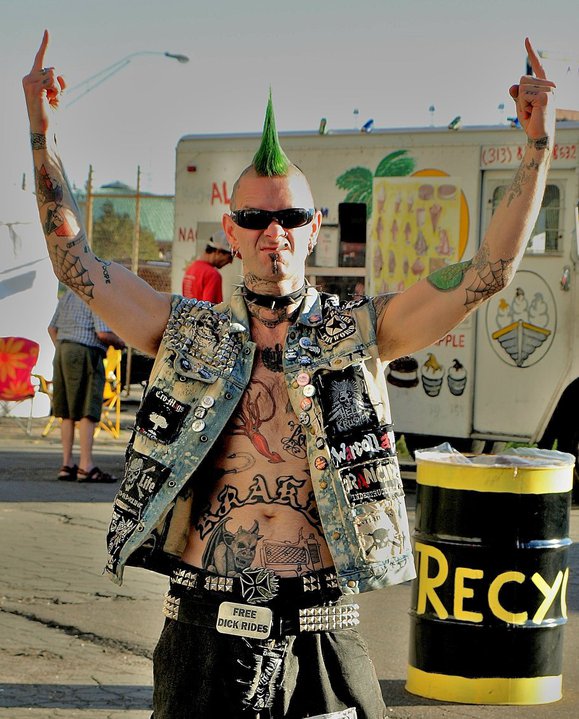 Front view of a person with a green mohawk, wearing punk rocker attire, with raised arms showing their middle fingers