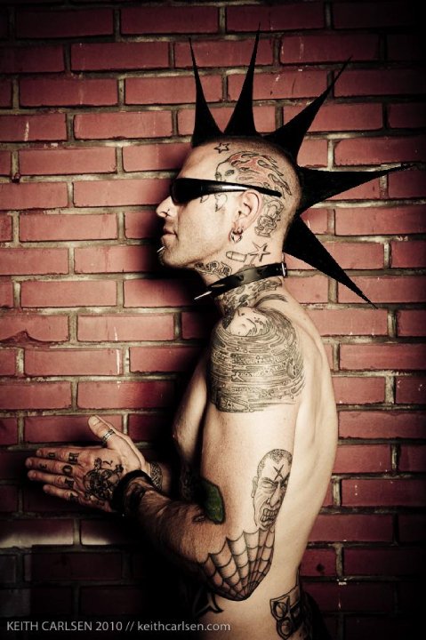 Left, waist up profile of a person with a spiked mohawk and body covered in tattoos, next to a brick wall