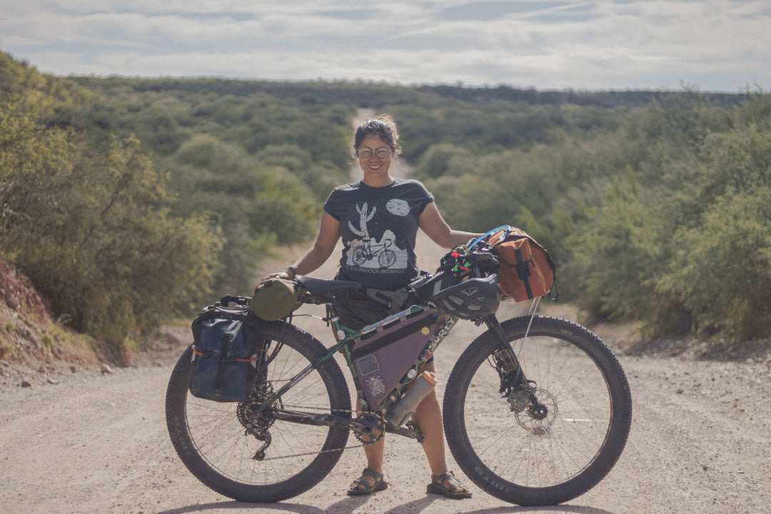 Cyclist on a fat bike with gear looks up a hill from a desert gravel road with large cactuses and hills in background