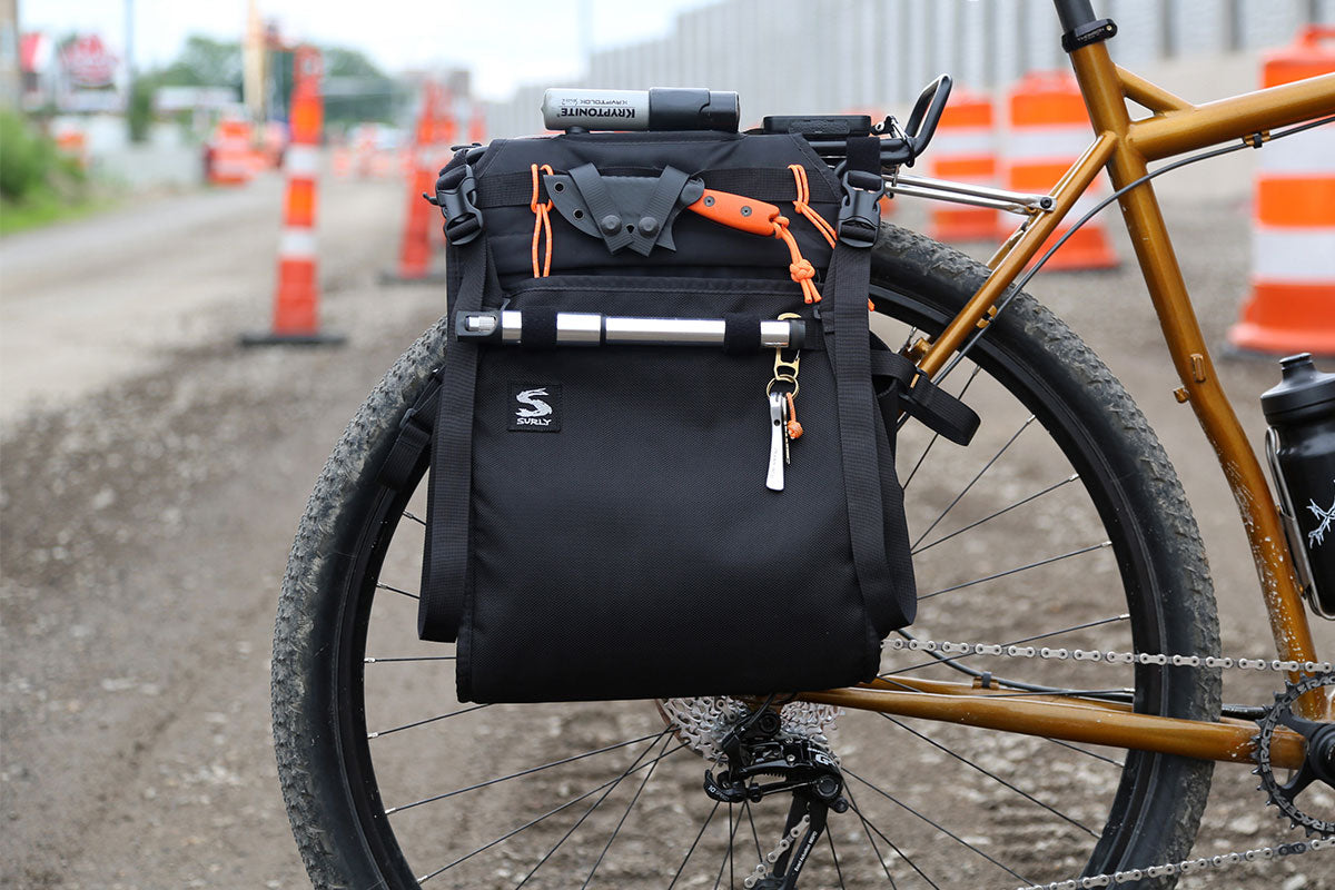 A black bicycle pannier bag with orange accents and a silver handlebar mount, attached to the rear rack of a gold-framed bicycle on a dirt road with orange traffic cones in the background.