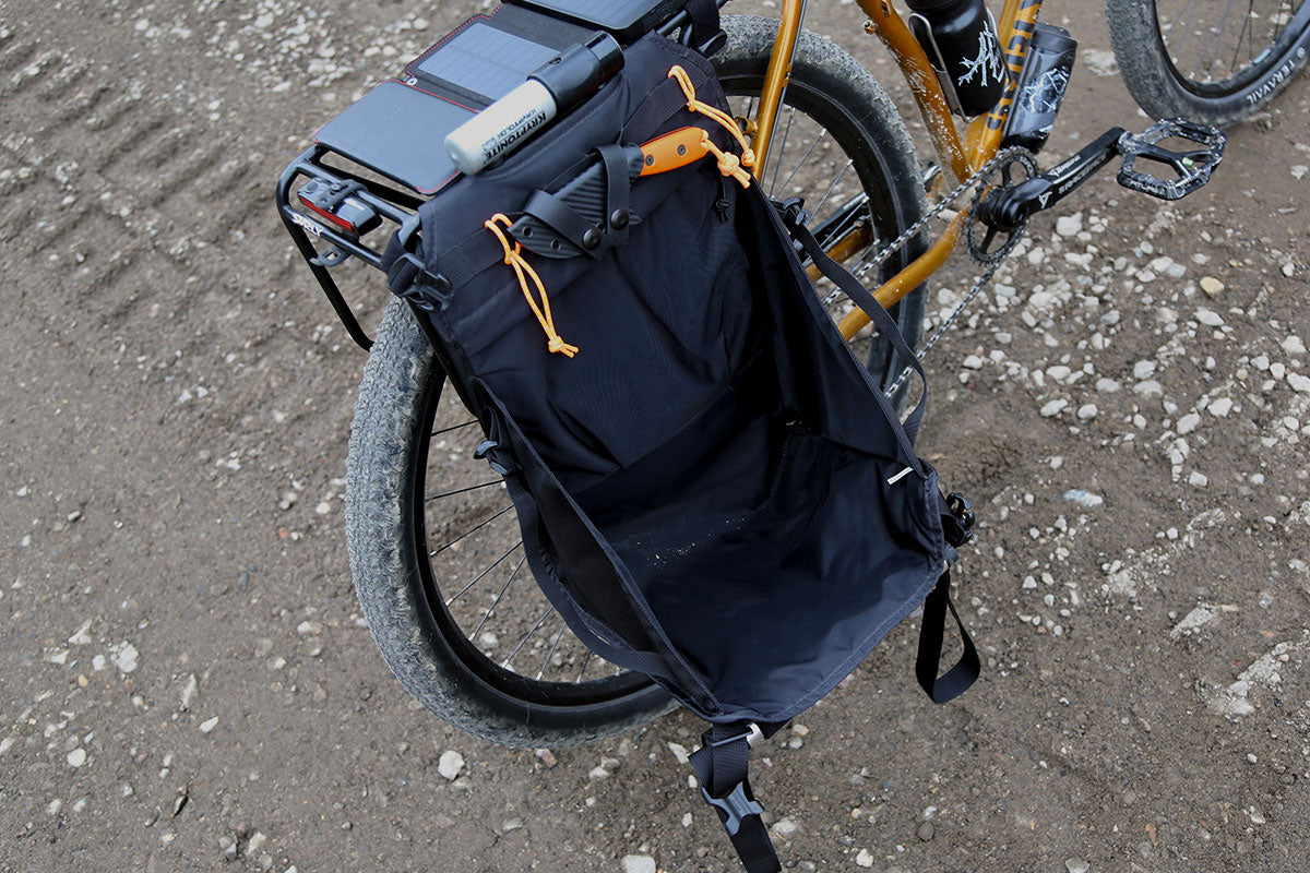 A black bicycle pannier bag with orange drawstring accents and a black handlebar bag, mounted on a yellow bicycle on a dirt and gravel path.