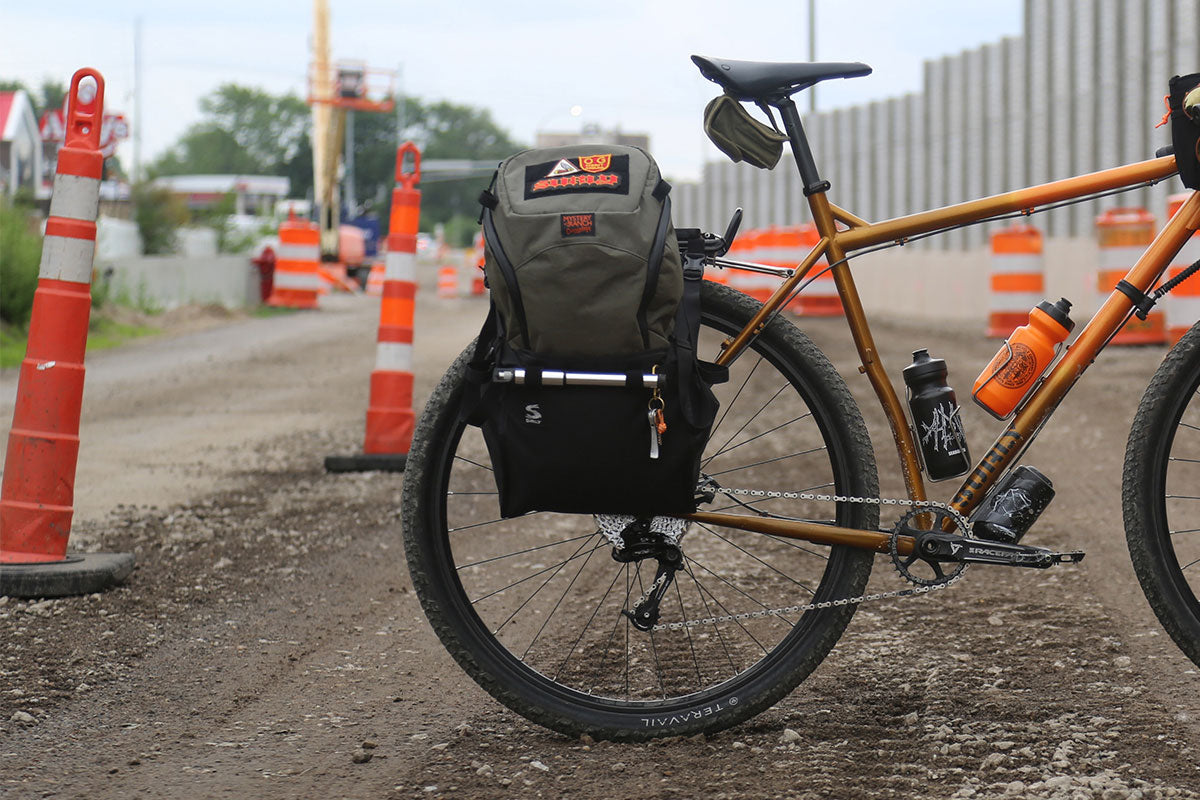 A rear pannier bag mounted on the back of an orange bicycle, photographed outdoors on a dirt construction site with orange traffic cones in the background.