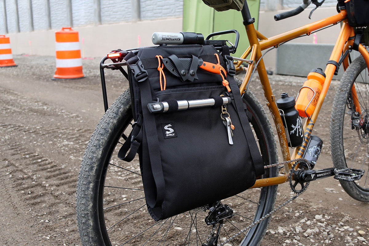 A black saddlebag bicycle pannier bag with orange accents and a silver tripod mount, attached to the rear rack of an orange bicycle on a dirt construction site.