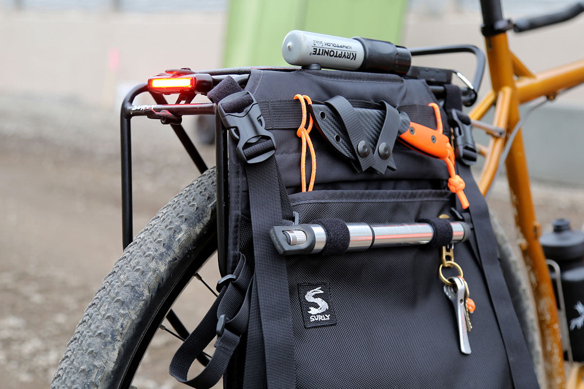 A black bicycle pannier bag mounted on the rear rack of a bicycle, shown outdoors. The bag has orange accents, a silver bicycle pump, a small flashlight, and a keychain, with a visible 'S' logo patch on the front pocket.