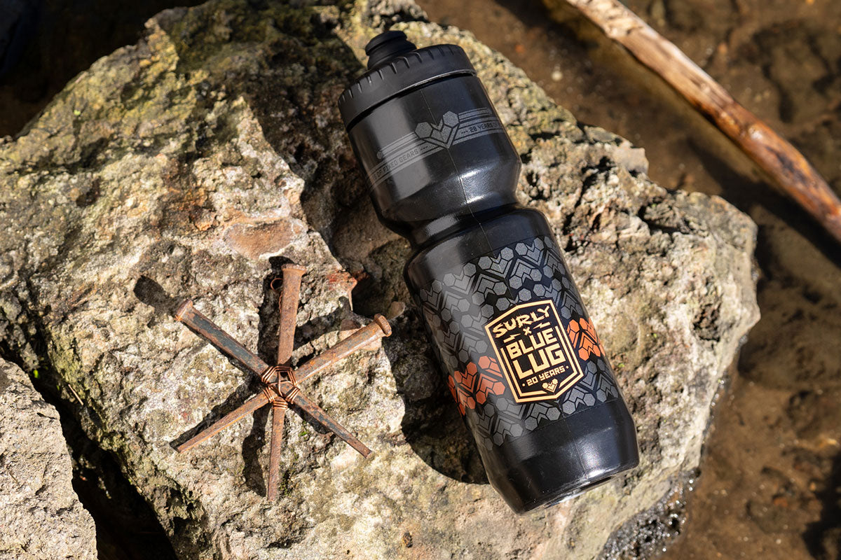 A black Surly Blue Lug water bottle with orange and gold graphics, resting on a textured gray rock outdoors next to rusty nails.