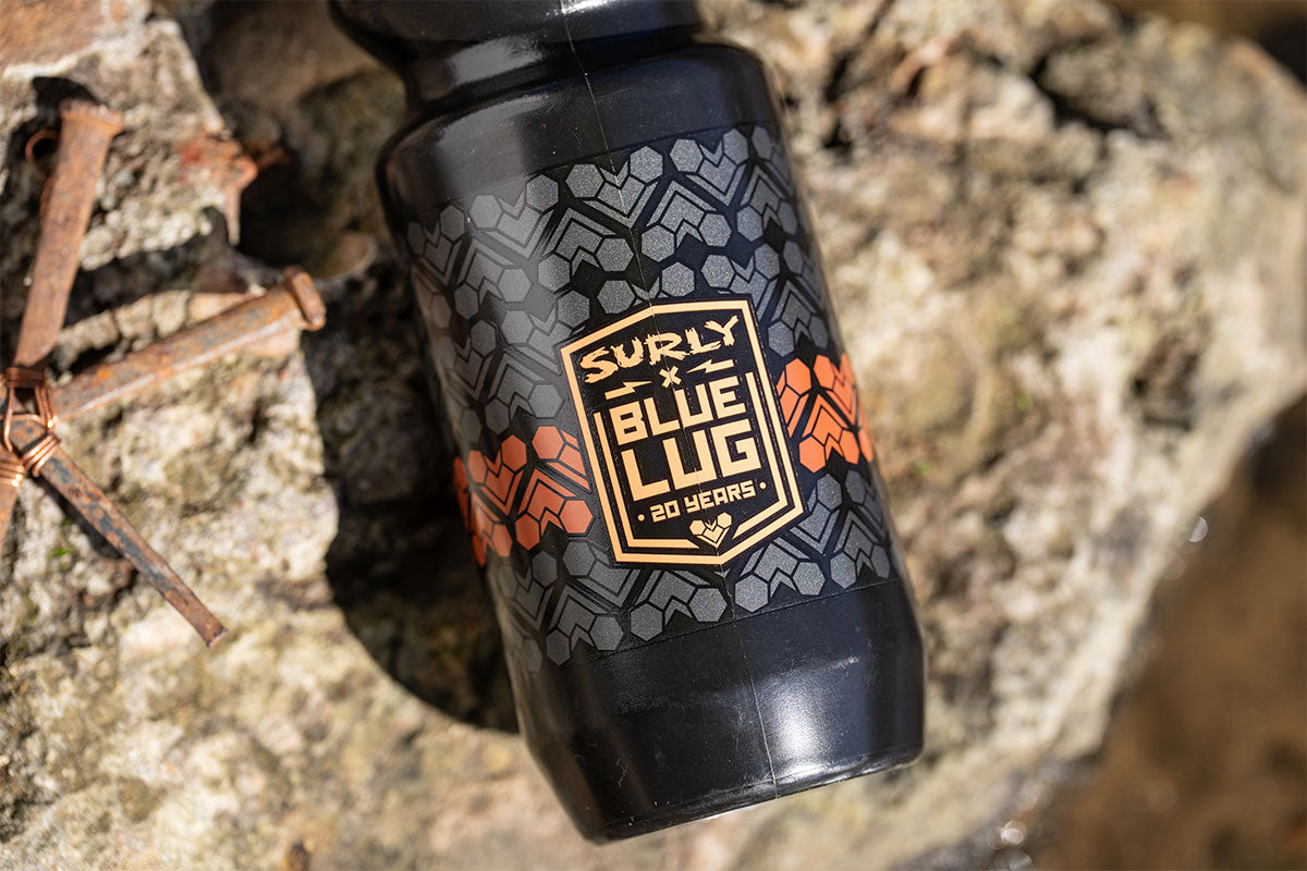 A black bike water bottle with a gray geometric pattern and orange accents, featuring a prominent 'SURLY x BLUE LUG + 20 YEARS +' shield logo, resting on a textured natural rock surface.