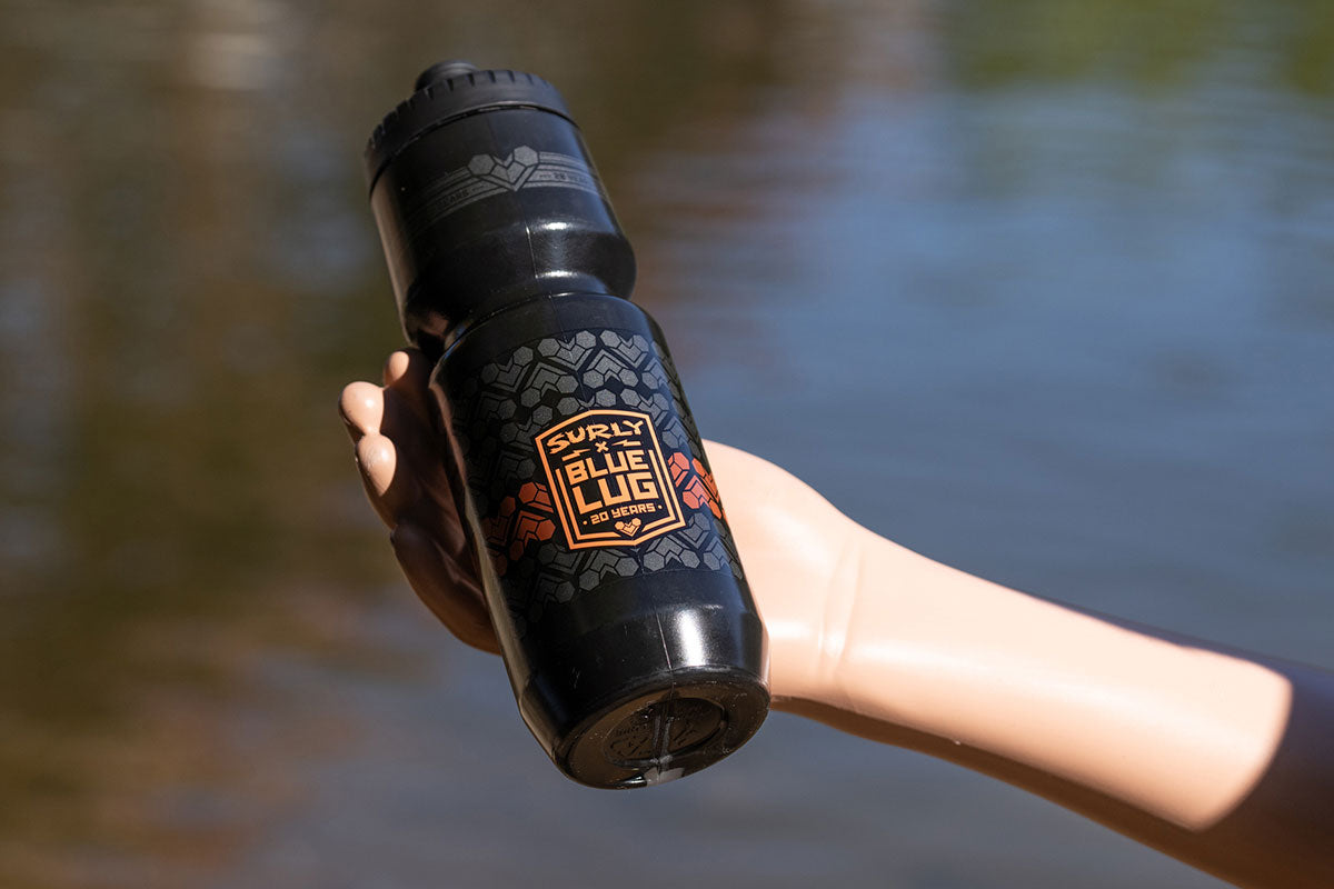 A black plastic water bottle with a black lid, featuring a graphic label that reads 'SURLY x BLUE LUG' in orange and white, held by a person's hand against a blurred outdoor water background.