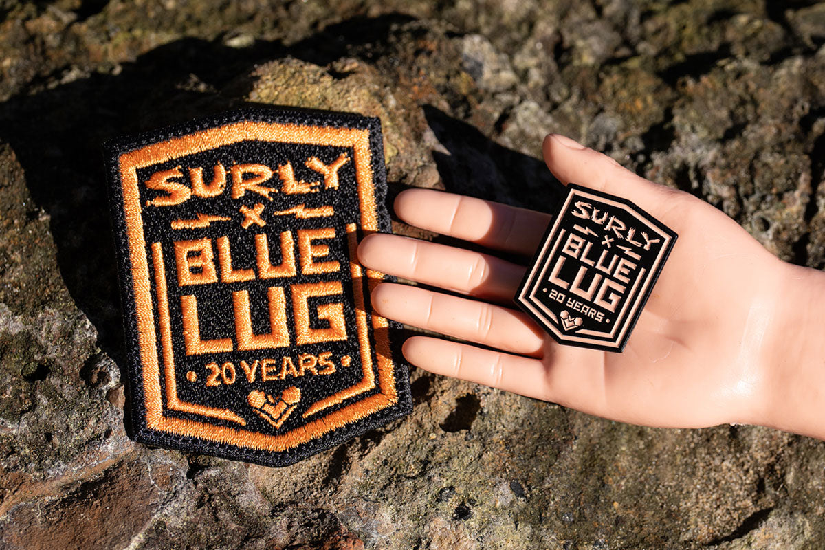 Surly x Blue Lug Patch and matching pin outdoors in natural sun on a rock with hand holding pin