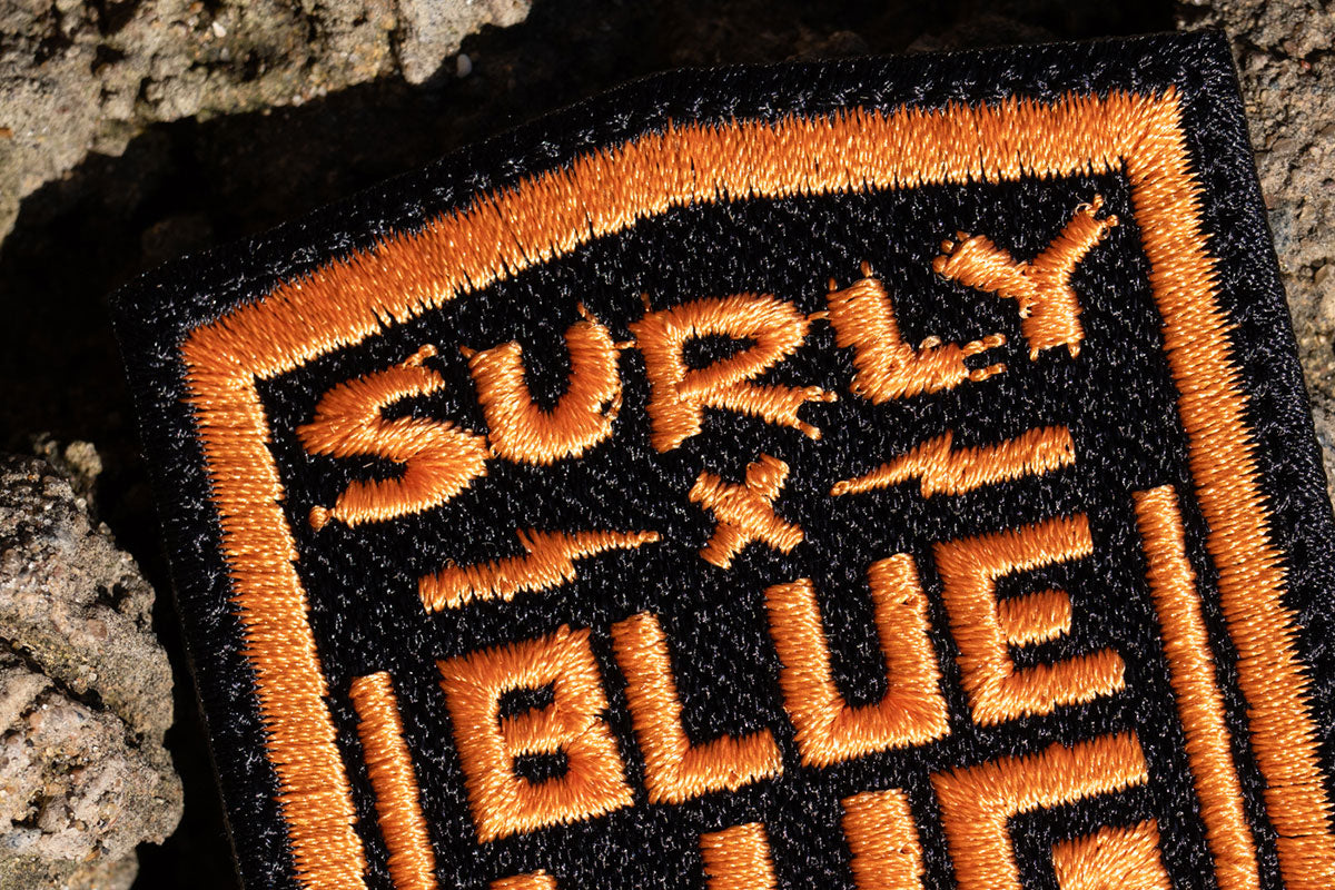 Close-up of an embroidered patch with orange lettering reading 'SURLY & BLUE' on a black background, placed against rough stone.