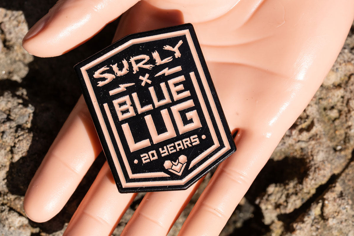 A close-up of a hand holding a shield-shaped pin that reads 'SURLY x BLUE LUG 20 YEARS' in light beige lettering on a black background, with rocky ground blurred in the background.