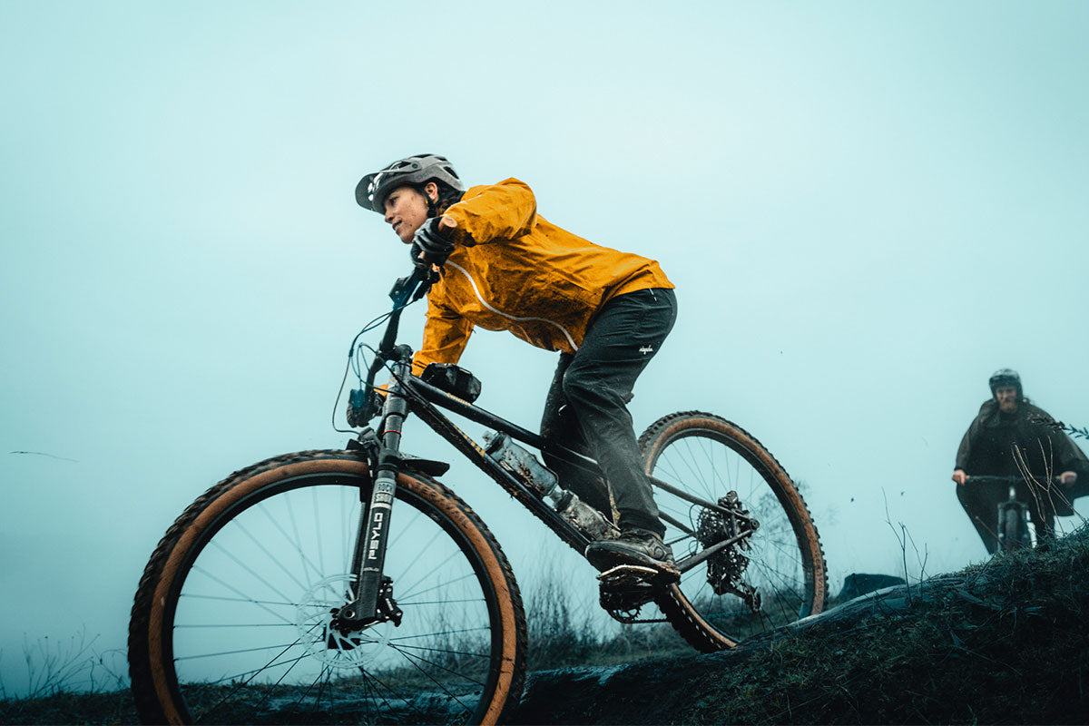 Person riding a mountain bike on a trail with another rider in the background.