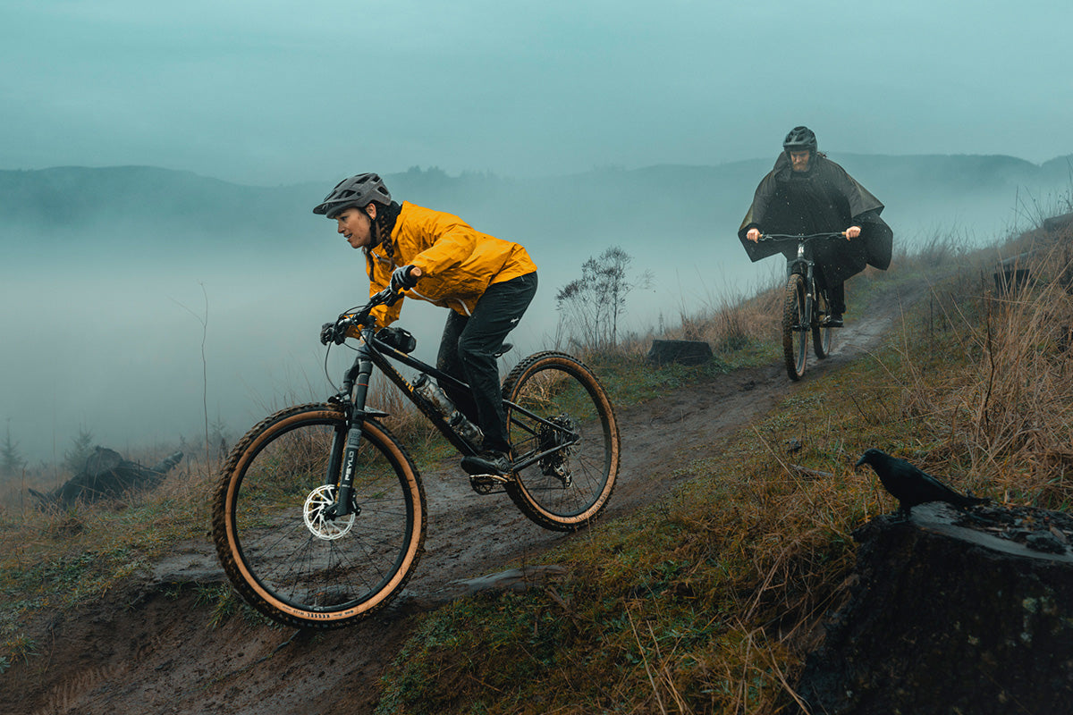 Two mountain bikers riding on a trail with a misty landscape in the background.
