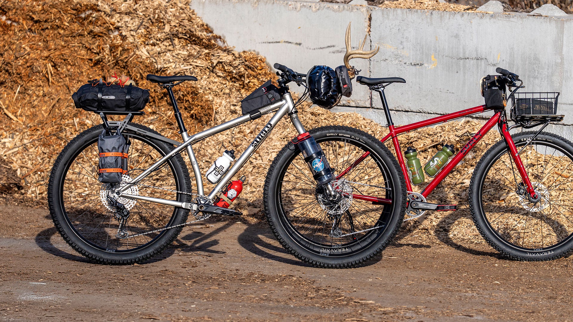 Two bicycles, one silver Krampus with bags, and one red Bridge Club, parked next to wall with wood chips in the background.