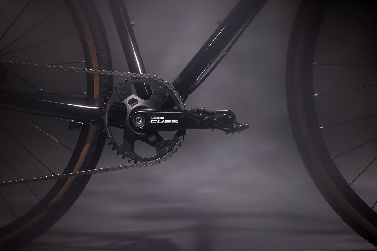 Close-up of a bicycle chain and gear system with 'CUES' branding on a dark background.